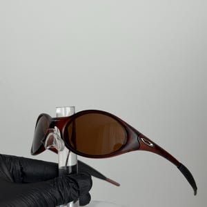 May include: Brown-tinted sunglasses with a dark red frame. The sunglasses have a curved design and are held by a clear display stand. The lenses are oval-shaped and the arms curve down.
