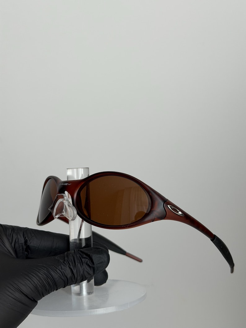 May include: Brown-tinted sunglasses with a dark red frame. The sunglasses have a curved design and are held by a clear display stand. The lenses are oval-shaped and the arms curve down.
