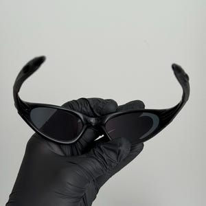May include: Black sunglasses with a curved frame and dark lenses. The sunglasses are held by a gloved hand against a plain white background. The sunglasses have a sporty, modern design.