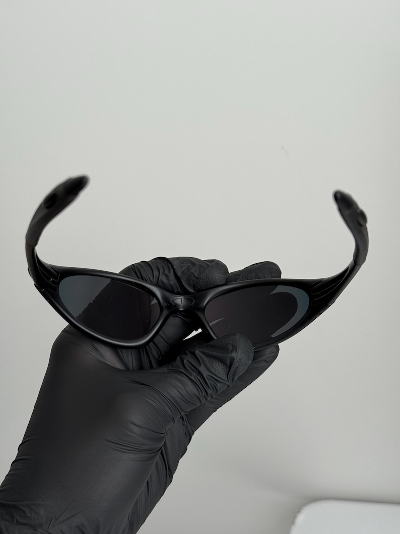 May include: Black sunglasses with a curved frame and dark lenses. The sunglasses are held by a gloved hand against a plain white background. The sunglasses have a sporty, modern design.
