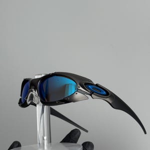 May include: Black sunglasses with blue mirrored lenses. The sunglasses have a sleek, modern design with a black frame and blue accents. The lenses reflect a bright blue color. The sunglasses are displayed on a clear stand.