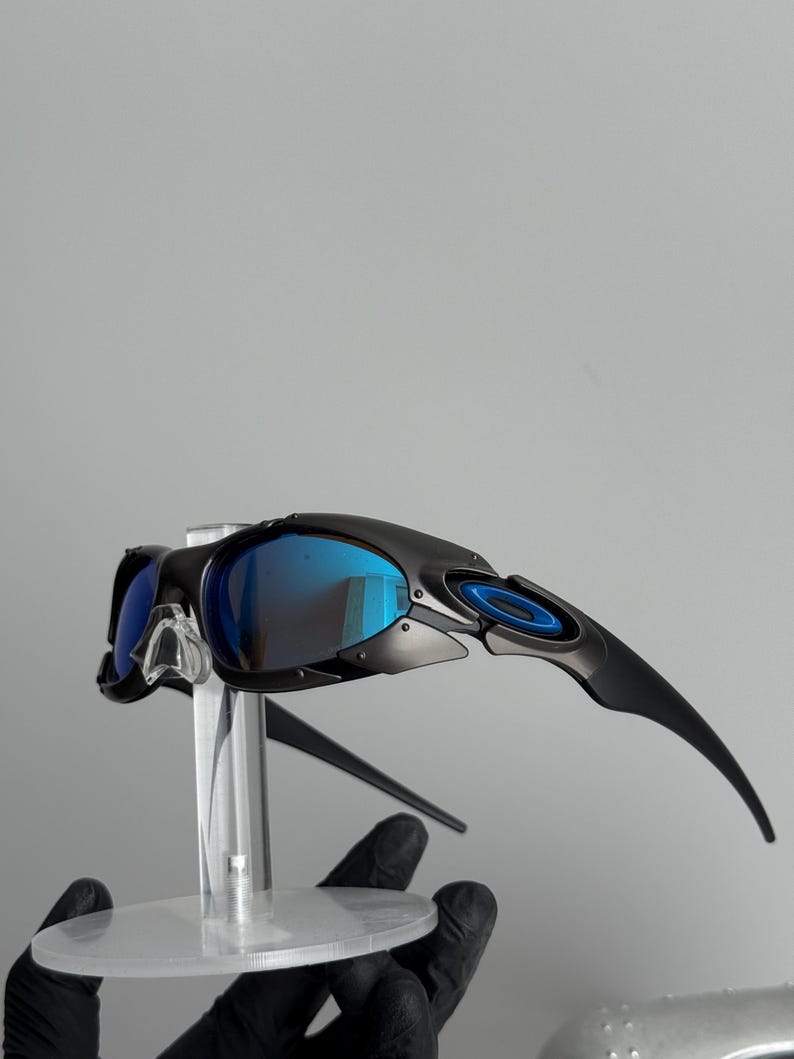 May include: Black sunglasses with blue mirrored lenses. The sunglasses have a sleek, modern design with a black frame and blue accents. The lenses reflect a bright blue color. The sunglasses are displayed on a clear stand.