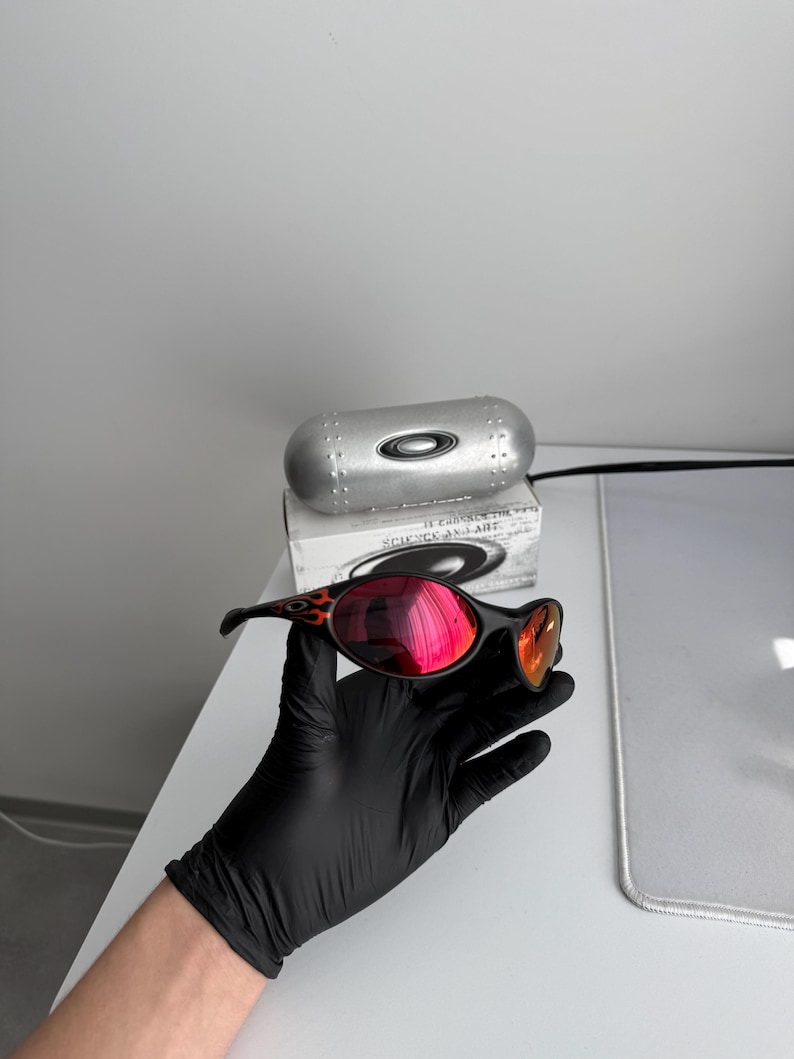 May include: Black sunglasses with red mirrored lenses and a flame design on the temples. The sunglasses are held in a black gloved hand. A silver case and box are in the background.