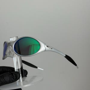 May include: Silver sunglasses with green lenses. The glasses have a modern design with curved arms and a small logo. The lenses reflect a green colour, and the frame is a metallic silver colour. The sunglasses are displayed on a clear stand.
