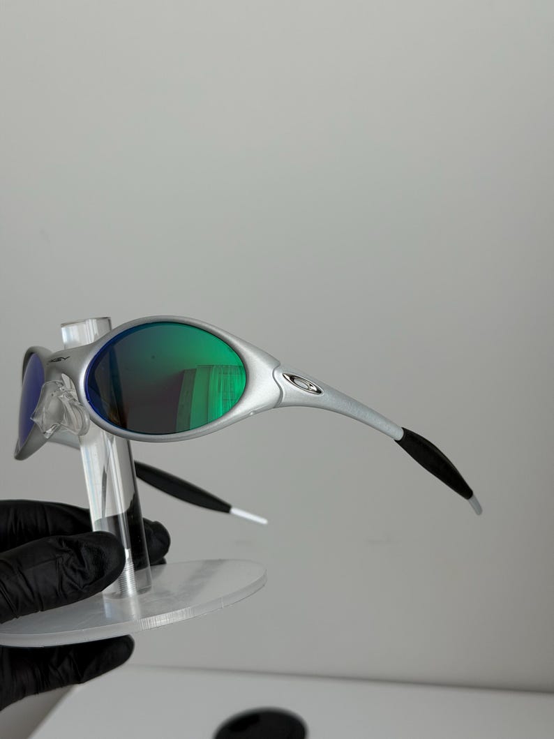 May include: Silver sunglasses with green lenses. The glasses have a modern design with curved arms and a small logo. The lenses reflect a green colour, and the frame is a metallic silver colour. The sunglasses are displayed on a clear stand.