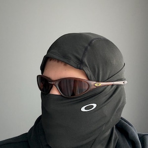 May include: Brown-framed sunglasses with a dark lens are worn over a black balaclava. The balaclava has a white logo on the front. The sunglasses have a gold accent on the temple.