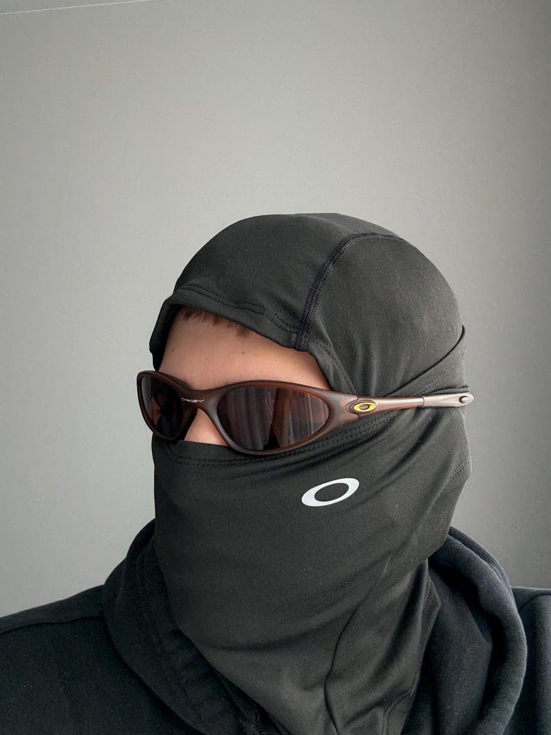 May include: Brown-framed sunglasses with a dark lens are worn over a black balaclava. The balaclava has a white logo on the front. The sunglasses have a gold accent on the temple.
