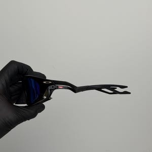 May include: Black sunglasses with blue lenses. The sunglasses have a sleek, modern design with a black frame and arms. The lenses are a vibrant blue color, and the overall look is sporty and stylish.