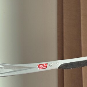 May include: Close-up of a silver-coloured eyeglass frame. The frame has a red and white label that reads "USA Assembled". The frame also has a CE marking.