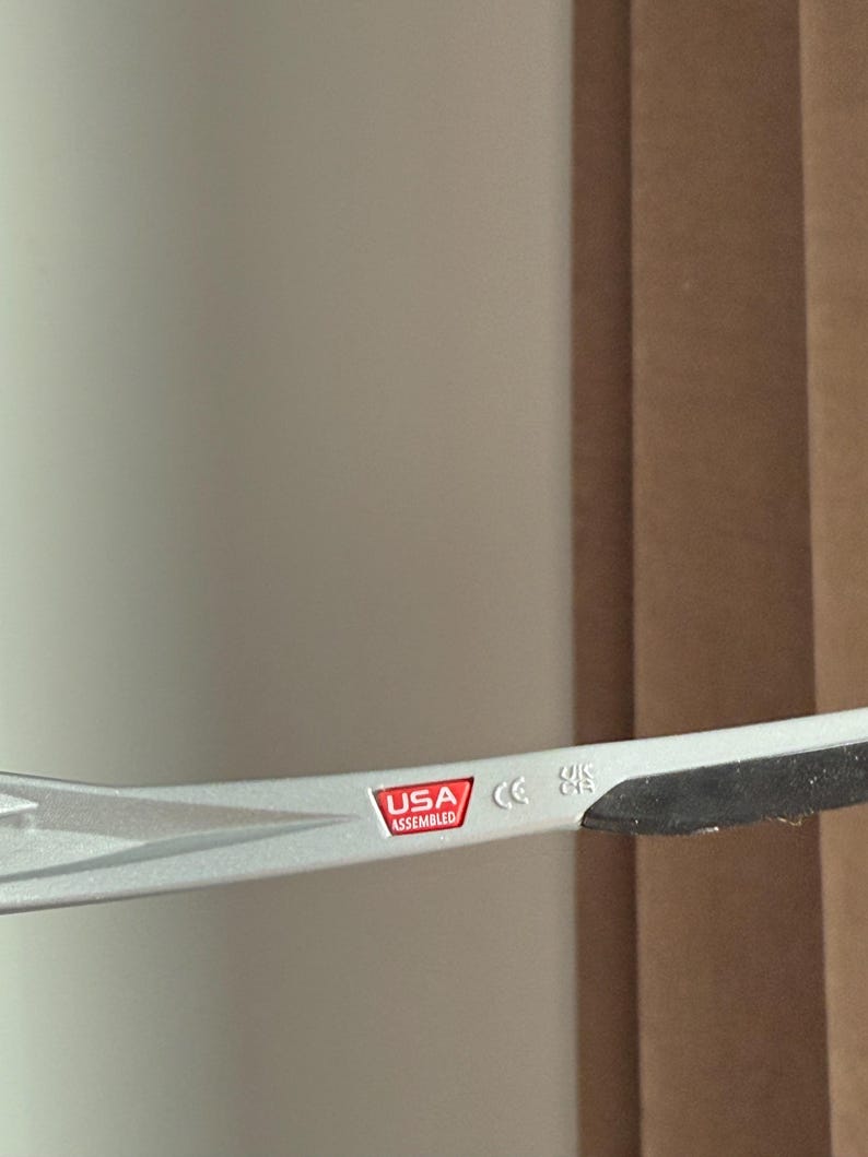 May include: Close-up of a silver-coloured eyeglass frame. The frame has a red and white label that reads "USA Assembled". The frame also has a CE marking.