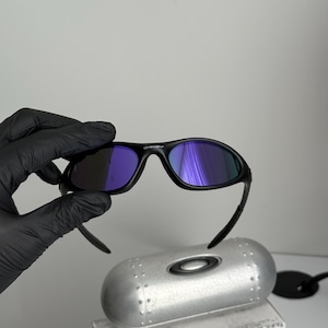 May include: Black sunglasses with purple-tinted lenses. The sunglasses are held above a silver case and a box with the text "SCIENCE AND ART". The sunglasses have a curved frame and a sleek design.
