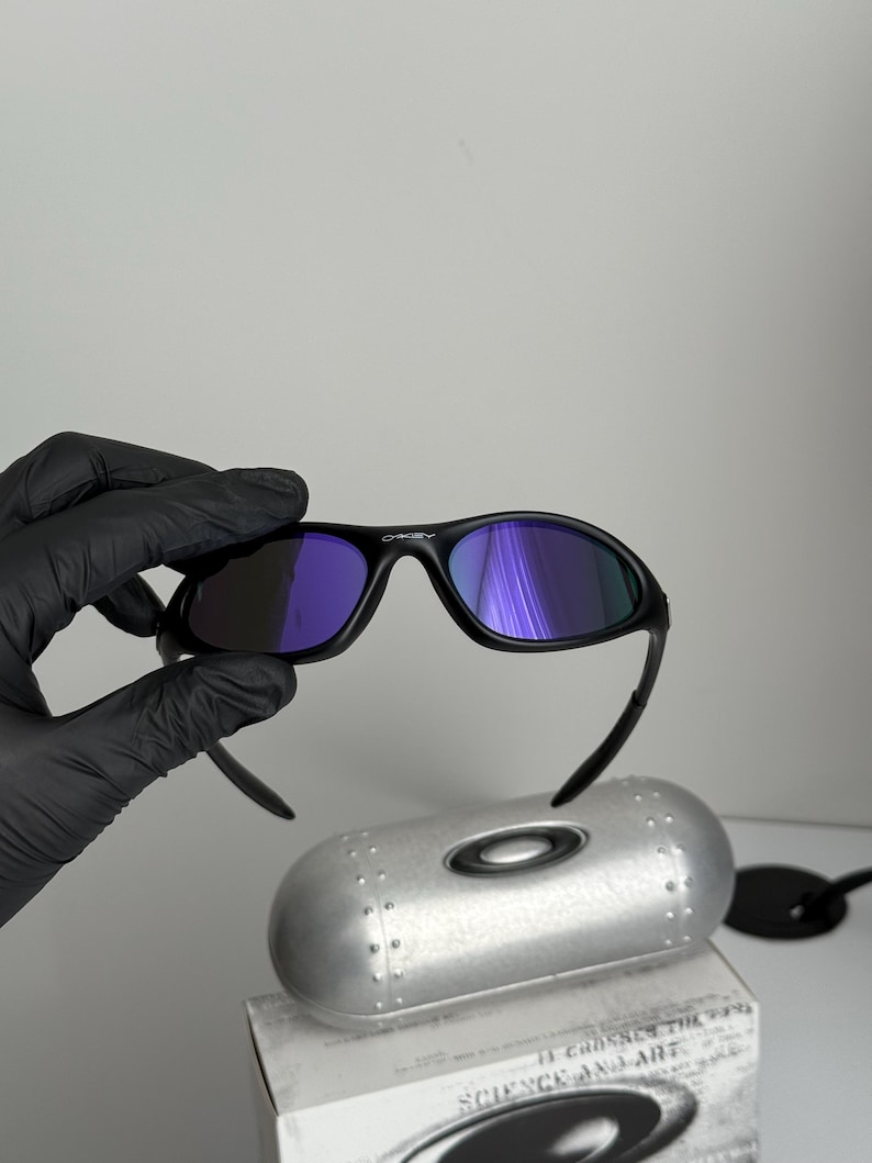 May include: Black sunglasses with purple-tinted lenses. The sunglasses are held above a silver case and a box with the text "SCIENCE AND ART". The sunglasses have a curved frame and a sleek design.