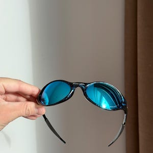 May include: Blue mirrored sunglasses with a black and blue speckled frame. The sunglasses have a curved design. The text "BRAIN DEAD" is visible on the top of the frame.