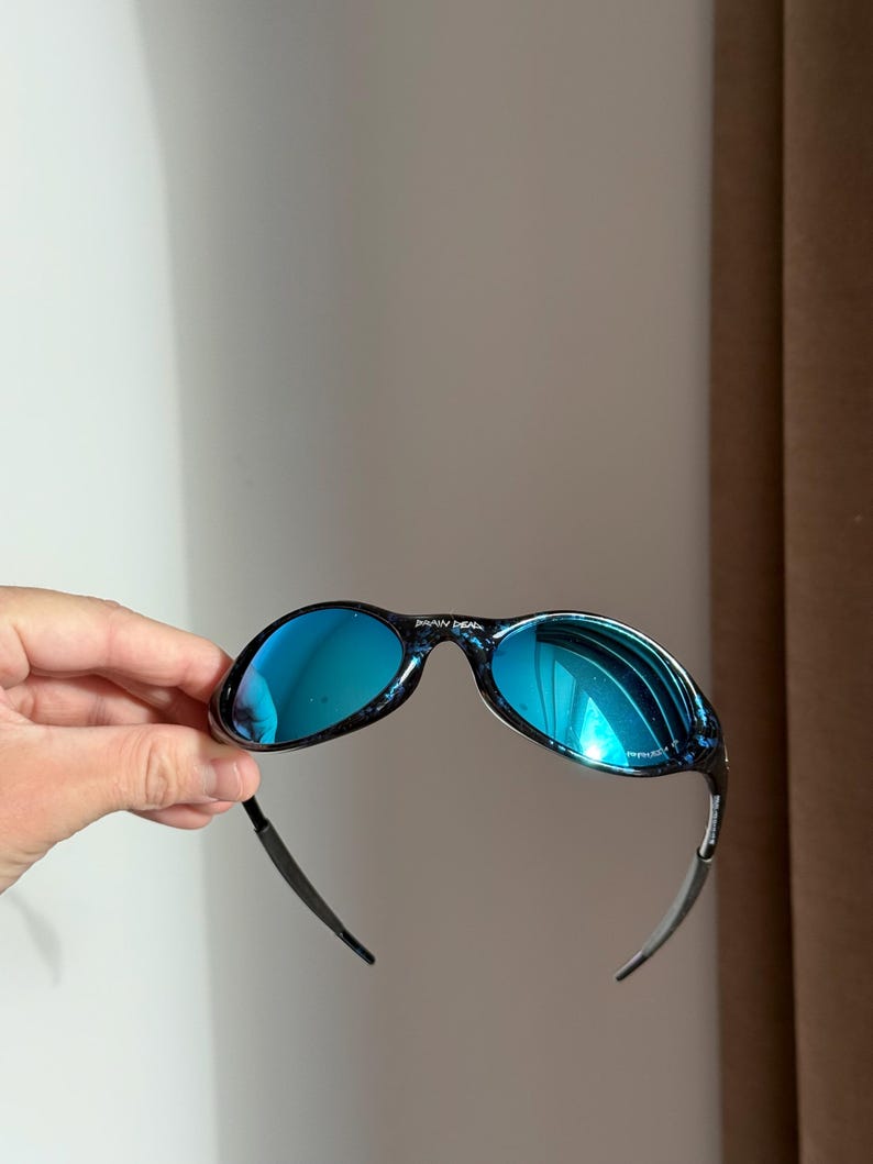 May include: Blue mirrored sunglasses with a black and blue speckled frame. The sunglasses have a curved design. The text "BRAIN DEAD" is visible on the top of the frame.