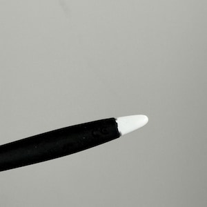 May include: A black stylus with a white tip. The stylus is angled towards the upper right corner against a gray background. The tip is a rounded, smooth shape.