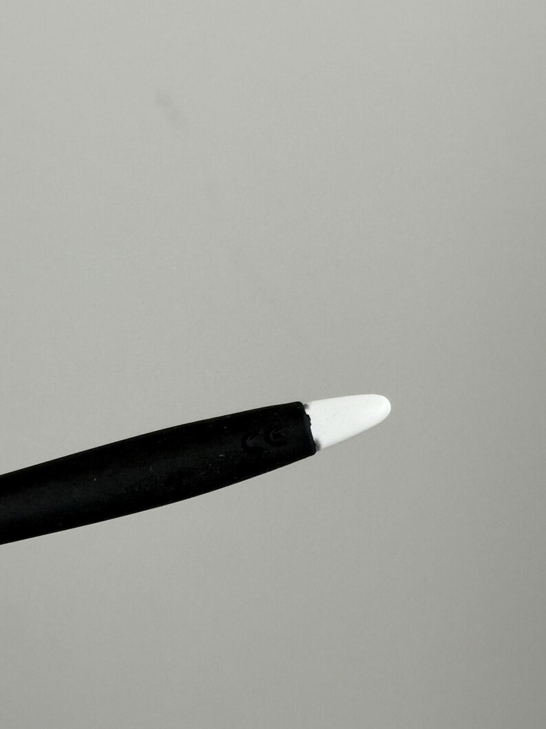 May include: A black stylus with a white tip. The stylus is angled towards the upper right corner against a gray background. The tip is a rounded, smooth shape.