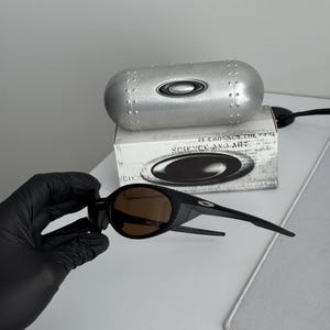 Puede incluir: Gafas de sol negras con lentes marrones y estuche plateado. El estuche tiene un dise&ntilde;o remachado y el logotipo de Oakley. La caja detr&aacute;s de las gafas de sol tiene el texto "SCIENCE AND ART".