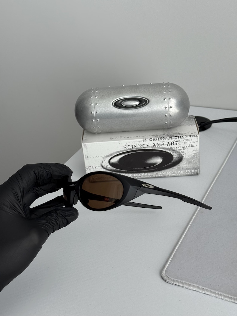 Puede incluir: Gafas de sol negras con lentes marrones y estuche plateado. El estuche tiene un dise&ntilde;o remachado y el logotipo de Oakley. La caja detr&aacute;s de las gafas de sol tiene el texto "SCIENCE AND ART".