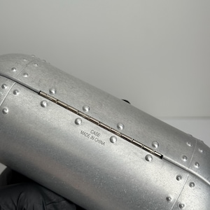 May include: Silver-colored, riveted case with a hinged opening. The case has a cylindrical shape and is made of a metallic material. The text "CASE MADE IN CHINA" is printed on the case.