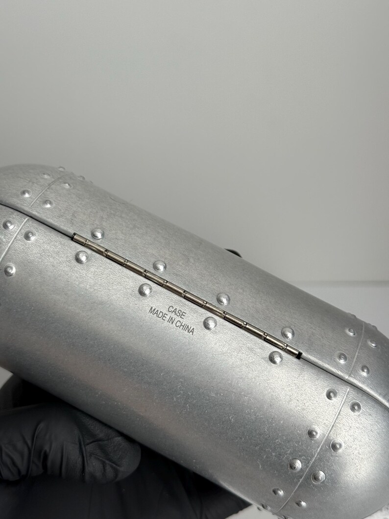 May include: Silver-colored, riveted case with a hinged opening. The case has a cylindrical shape and is made of a metallic material. The text "CASE MADE IN CHINA" is printed on the case.