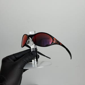 May include: Black sunglasses with red lenses and a flame design on the temples. The sunglasses are displayed on a clear stand. The lenses reflect the surrounding environment.