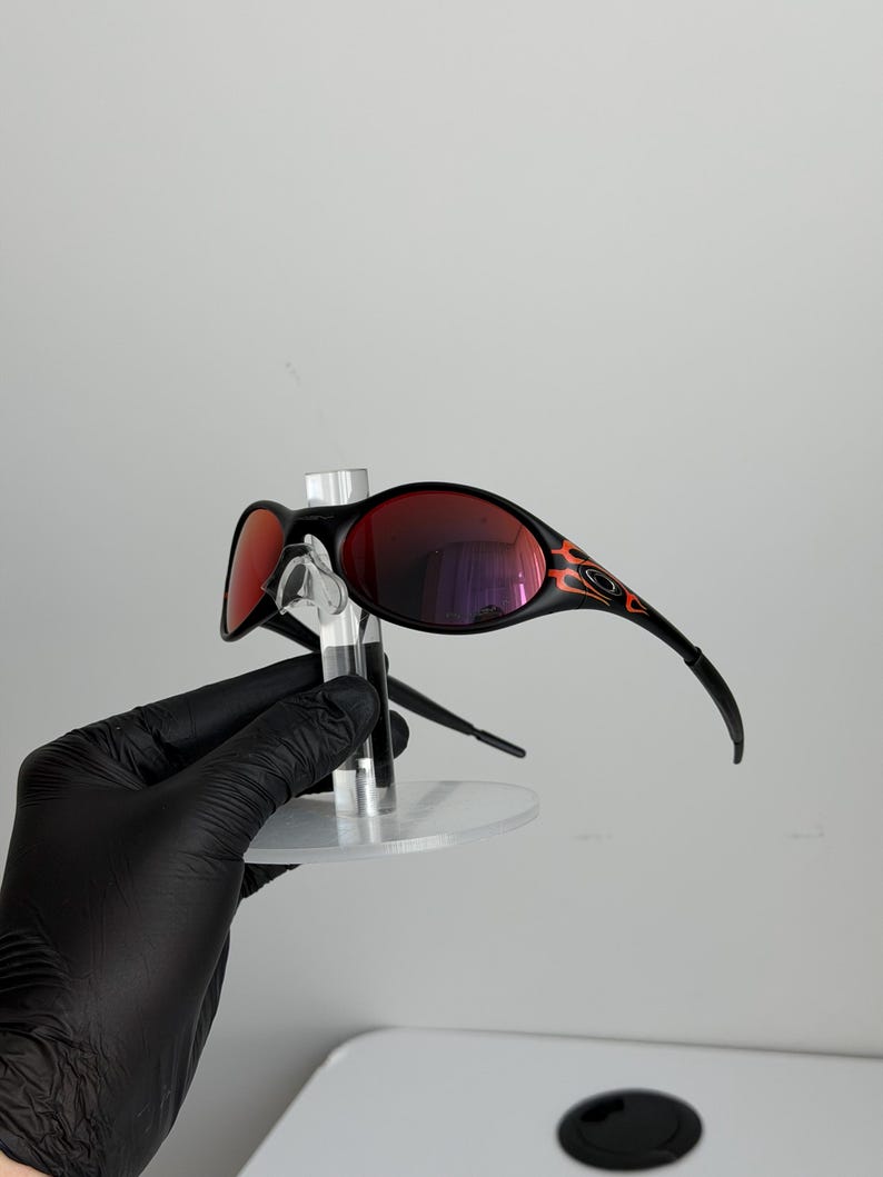May include: Black sunglasses with red lenses and a flame design on the temples. The sunglasses are displayed on a clear stand. The lenses reflect the surrounding environment.