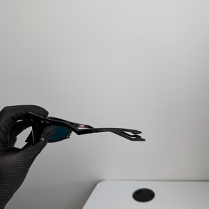 May include: Black sunglasses with a sleek, modern design. The sunglasses have dark lenses and a black frame. The arms of the sunglasses have a unique shape. The sunglasses are held by a gloved hand.
