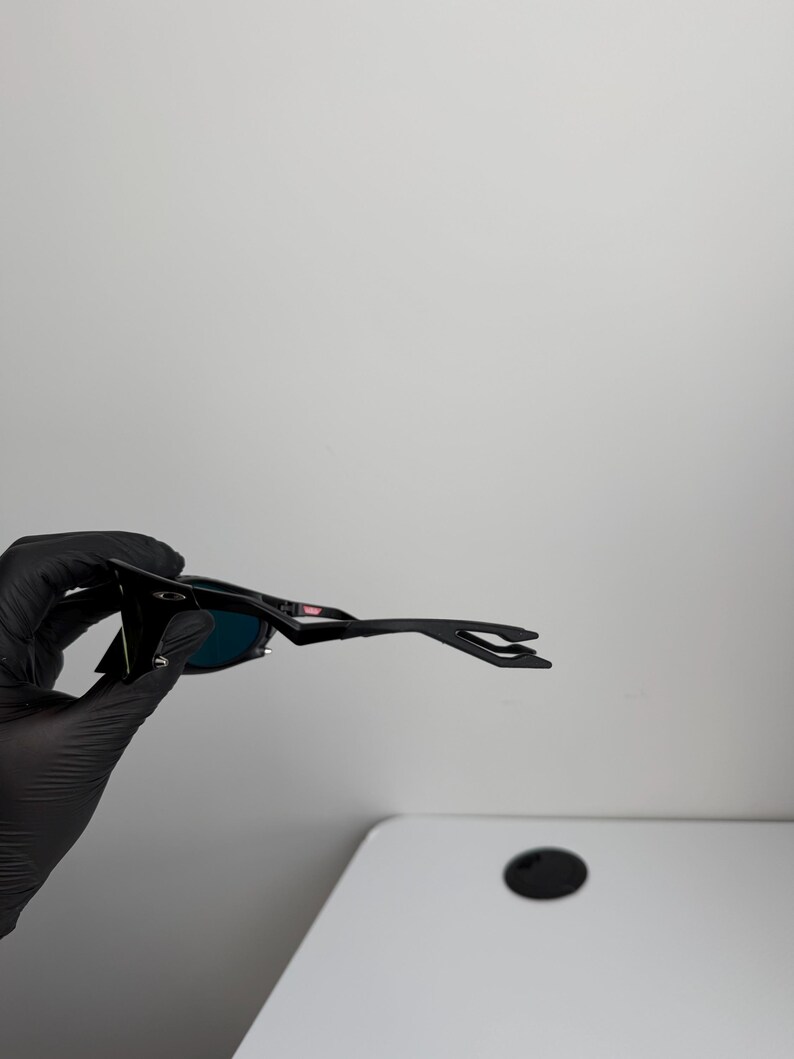 May include: Black sunglasses with a sleek, modern design. The sunglasses have dark lenses and a black frame. The arms of the sunglasses have a unique shape. The sunglasses are held by a gloved hand.