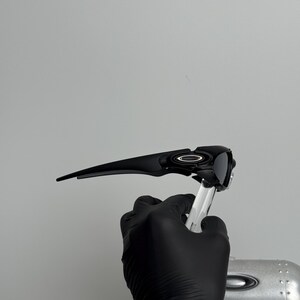 May include: Black sunglasses with a sleek, modern design. The sunglasses have a dark frame and lenses, with a silver emblem on the side. The sunglasses are held by a gloved hand.