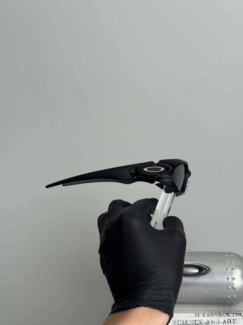 May include: Black sunglasses with a sleek, modern design. The sunglasses have a dark frame and lenses, with a silver emblem on the side. The sunglasses are held by a gloved hand.