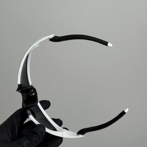 Puede incluir: Gafas de sol deportivas blancas y negras con un dise&ntilde;o elegante y moderno. Las monturas son blancas con detalles y lentes negros. Las patillas son negras con puntas blancas. Las gafas de sol est&aacute;n sujetas por una mano enguantada.