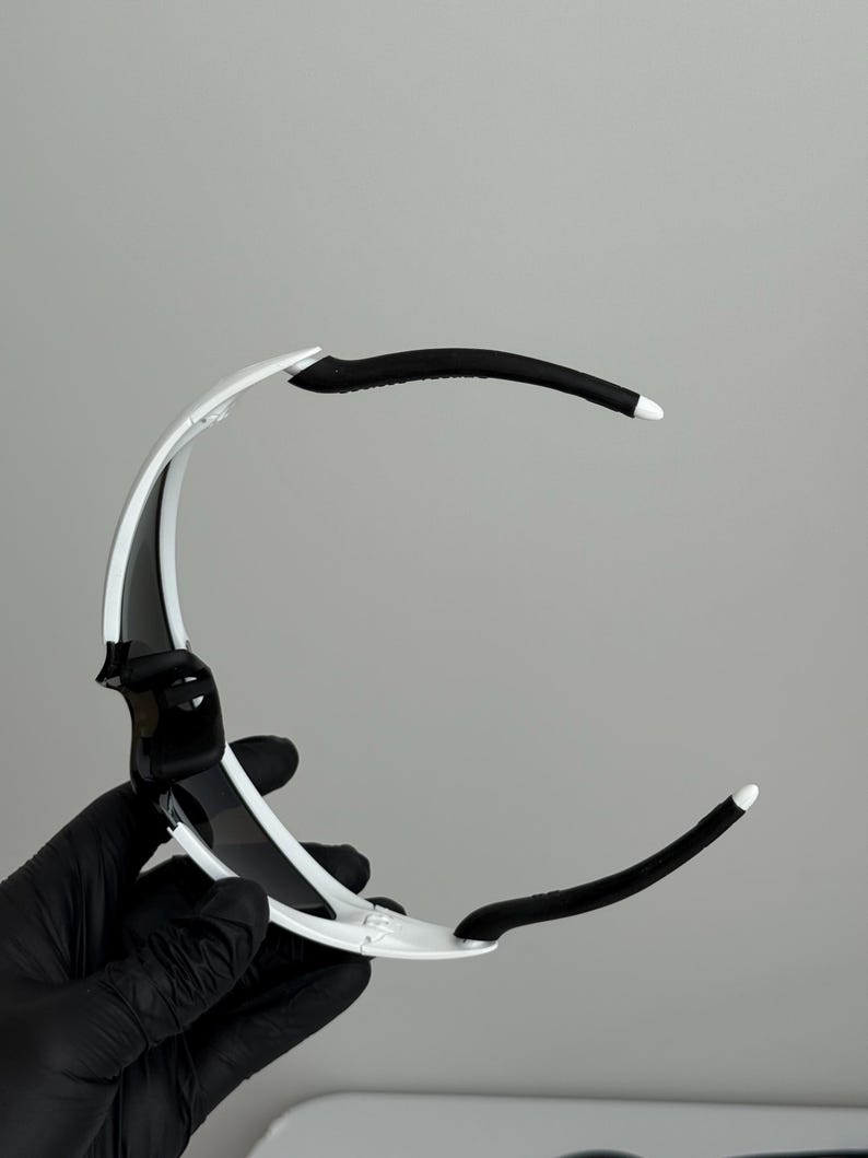 Puede incluir: Gafas de sol deportivas blancas y negras con un dise&ntilde;o elegante y moderno. Las monturas son blancas con detalles y lentes negros. Las patillas son negras con puntas blancas. Las gafas de sol est&aacute;n sujetas por una mano enguantada.