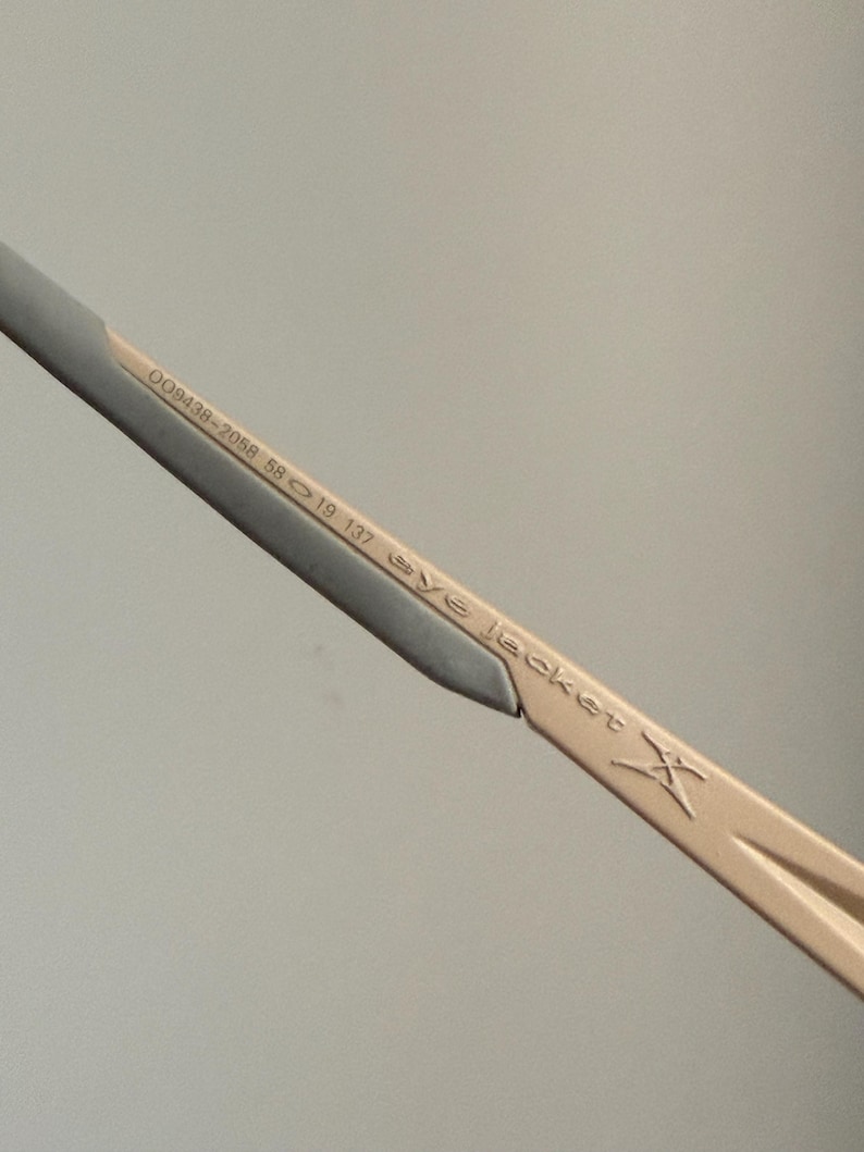 May include: Close-up of a portion of eyeglasses. The frame is a light tan color with a silver accent. The text "OO9438-2058 58 19 137 eye jacket X" is printed on the frame.