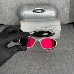 May include: Silver-framed sunglasses with bright red lenses are held by a gloved hand. The sunglasses are paired with a silver case and a white box with the text "SCIENCE AND ART".