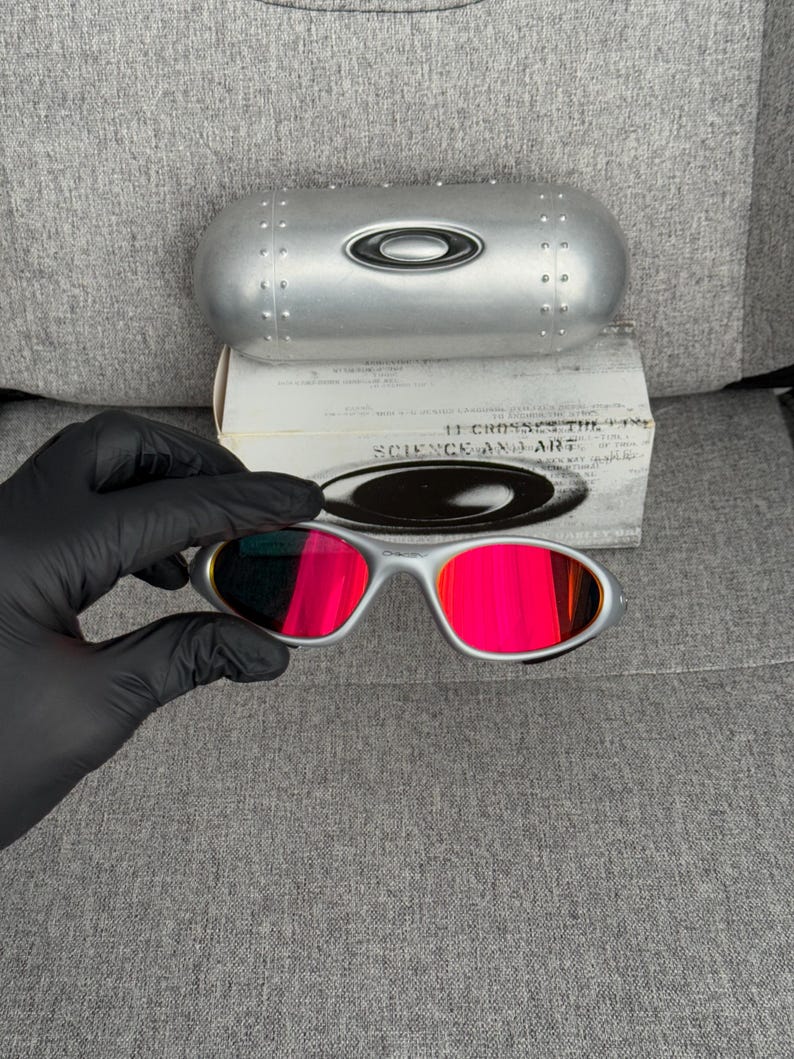 May include: Silver-framed sunglasses with bright red lenses are held by a gloved hand. The sunglasses are paired with a silver case and a white box with the text "SCIENCE AND ART".