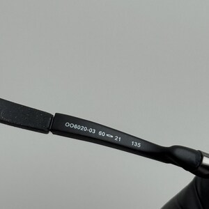 May include: Close-up of a black eyeglass frame arm with the numbers "OO6020-03 60-21 135" printed in white. The arm is slightly curved and has a matte finish.