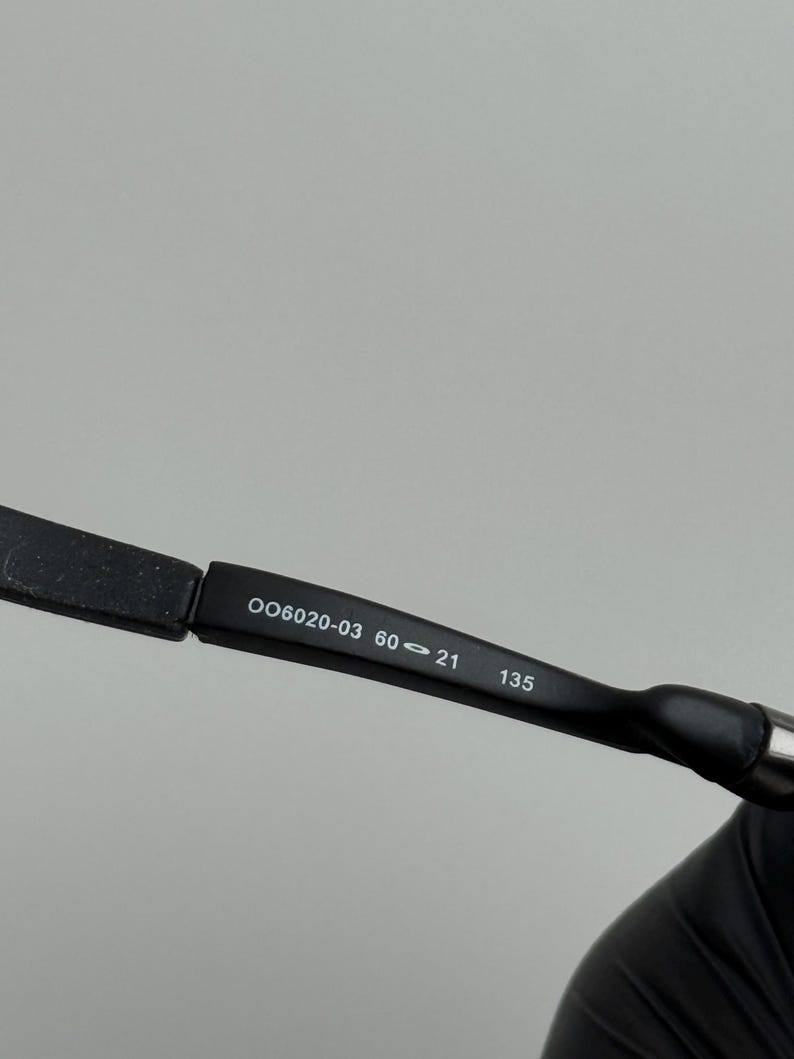 May include: Close-up of a black eyeglass frame arm with the numbers "OO6020-03 60-21 135" printed in white. The arm is slightly curved and has a matte finish.