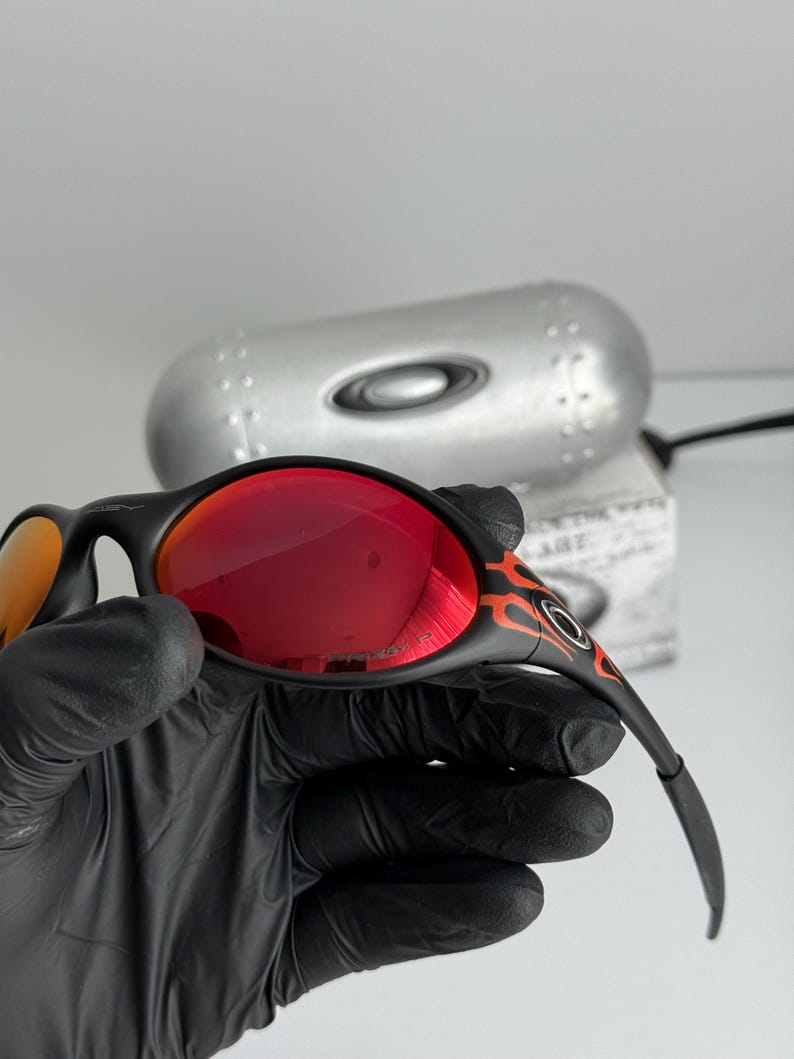 May include: Black sunglasses with red lenses and a flame design on the temples. The sunglasses are held by a gloved hand, with a silver case in the background. The lenses appear to be mirrored.