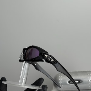 May include: Black sunglasses with purple lenses. The sunglasses have a sleek, modern design with a curved frame and a silver logo on the side. The lenses are a dark purple color, and the frame is black.