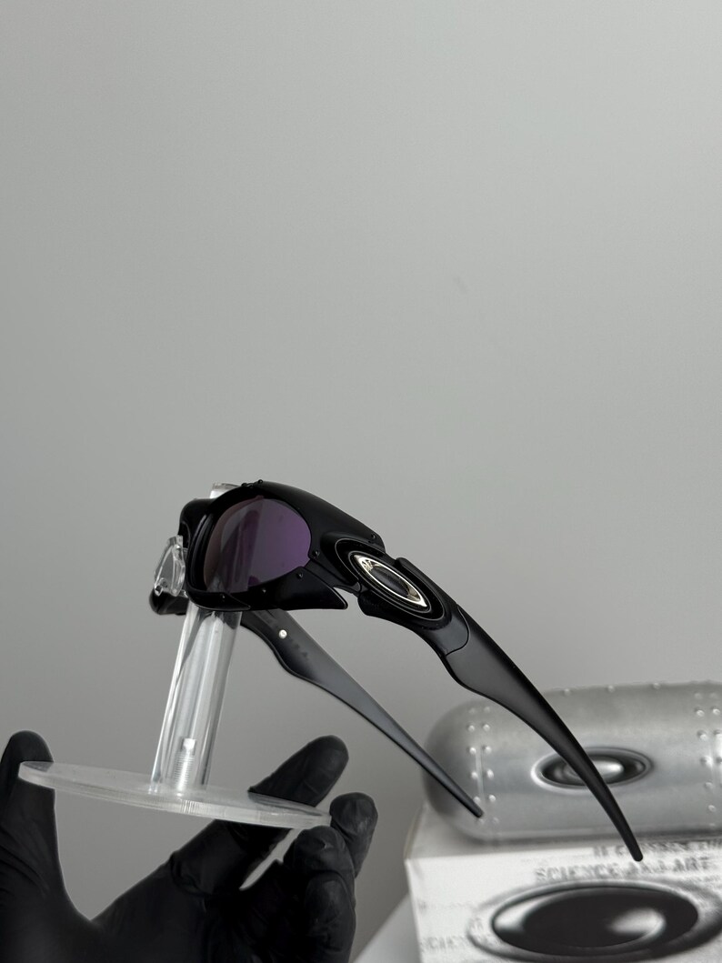 May include: Black sunglasses with purple lenses. The sunglasses have a sleek, modern design with a curved frame and a silver logo on the side. The lenses are a dark purple color, and the frame is black.