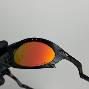May include: Black sunglasses with orange-red lenses. The lenses have the word "PRIZM" printed in white. The frame is black and has a sleek, modern design. The sunglasses are designed for sports and outdoor activities.