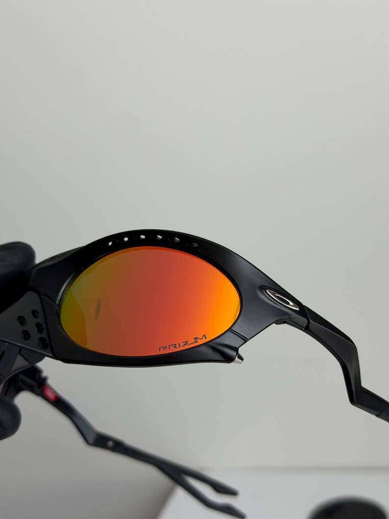May include: Black sunglasses with orange-red lenses. The lenses have the word "PRIZM" printed in white. The frame is black and has a sleek, modern design. The sunglasses are designed for sports and outdoor activities.