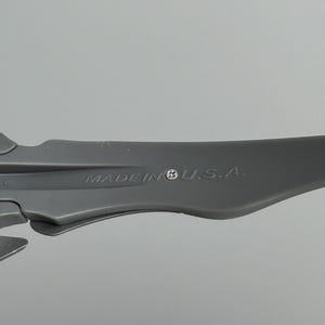 May include: A close-up shot of a gray plastic object with the text "MADE IN U.S.A." printed on it. The object has a curved, aerodynamic shape and appears to be a component of a larger product. The background is a plain, light gray.
