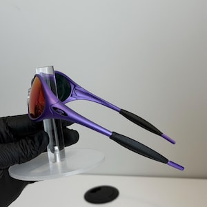 May include: Purple sunglasses with a red-orange lens. The sunglasses have a sleek, modern design with black accents on the arms. The glasses are displayed on a clear stand.