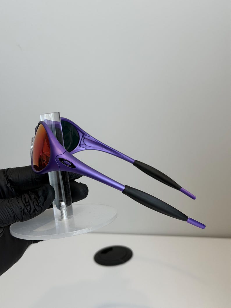 May include: Purple sunglasses with a red-orange lens. The sunglasses have a sleek, modern design with black accents on the arms. The glasses are displayed on a clear stand.