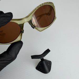 May include: Olive green sunglasses with brown lenses and a distinctive curved frame. A black stand is also visible, possibly for display or storage. The sunglasses' frame has a unique design.