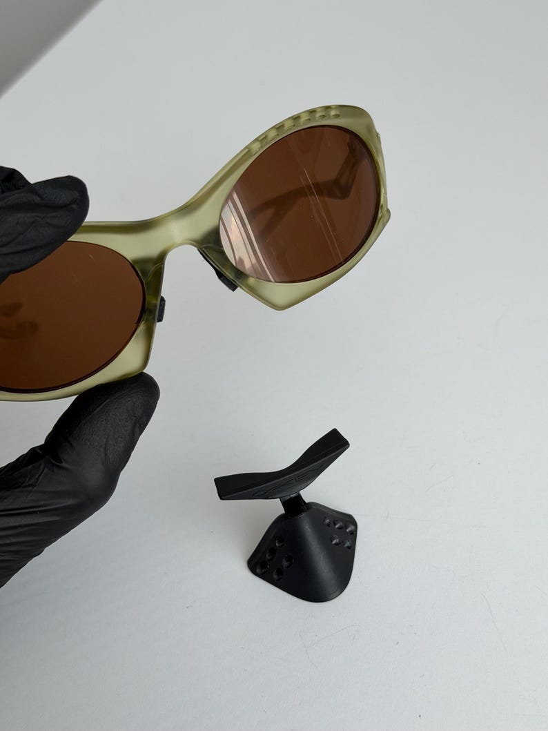May include: Olive green sunglasses with brown lenses and a distinctive curved frame. A black stand is also visible, possibly for display or storage. The sunglasses' frame has a unique design.