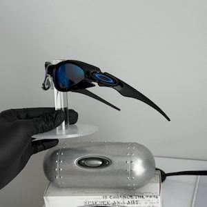 May include: Black sunglasses with blue lenses, displayed on a clear stand. The sunglasses have a sleek, modern design with a curved frame and the Oakley logo. A silver case and a box with text are also visible.