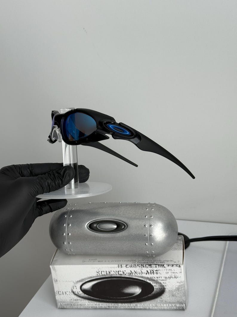 May include: Black sunglasses with blue lenses, displayed on a clear stand. The sunglasses have a sleek, modern design with a curved frame and the Oakley logo. A silver case and a box with text are also visible.