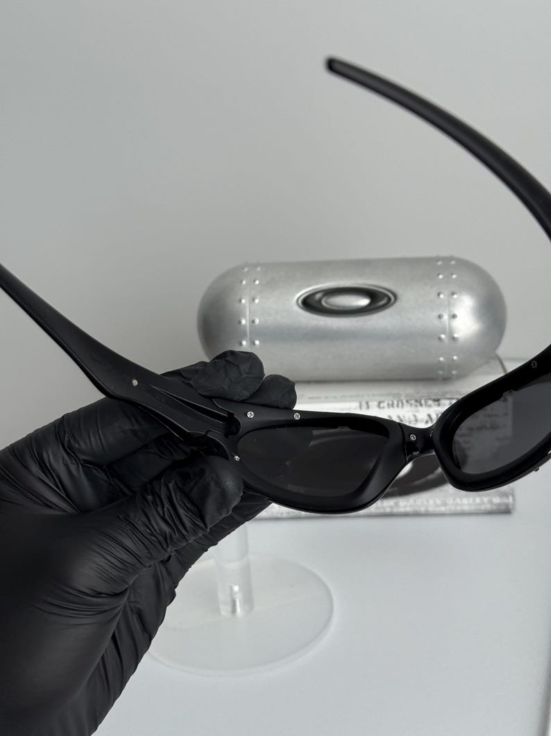 May include: Black sunglasses with a sleek, modern design. The sunglasses have a rectangular frame and dark lenses. A silver case with the Oakley logo is in the background.